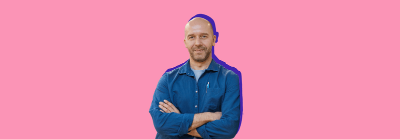 A smiling bald man in a denim shirt with his arms crossed, set against a pink background.