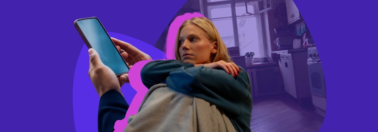 Young woman curled up on a sofa in a dim kitchen, holding up a phone with a blank screen