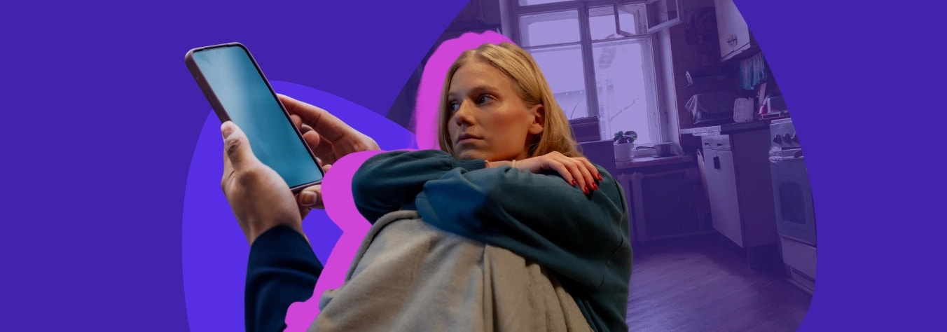 Young woman curled up on a sofa in a dim kitchen, holding up a phone with a blank screen