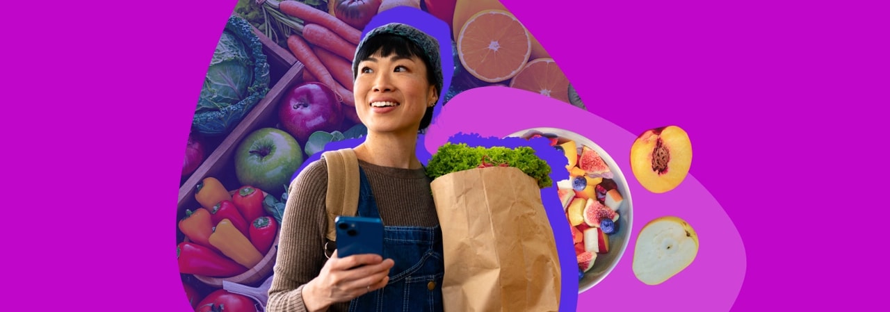 Smiling woman in dungarees holding a phone and a paper grocery bag, with a crate of fresh vegetables and a fruit bowl behind