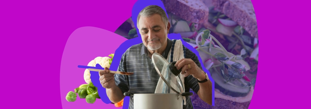 Older man smiling as he lifts the lid off a cooking pot, with fresh vegetables and a salad sandwich behind him