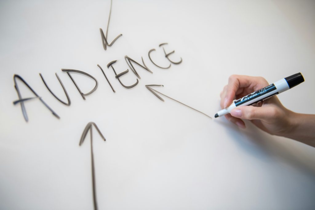 A hand holding a marker and writing the word “AUDIENCE” on a whiteboard with arrows pointing toward it, representing audience targeting, customer focus, marketing strategy, or consumer research.