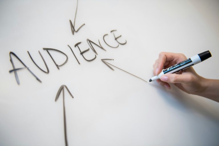 A hand holding a marker and writing the word “AUDIENCE” on a whiteboard with arrows pointing toward it, representing audience targeting, customer focus, marketing strategy, or consumer research.
